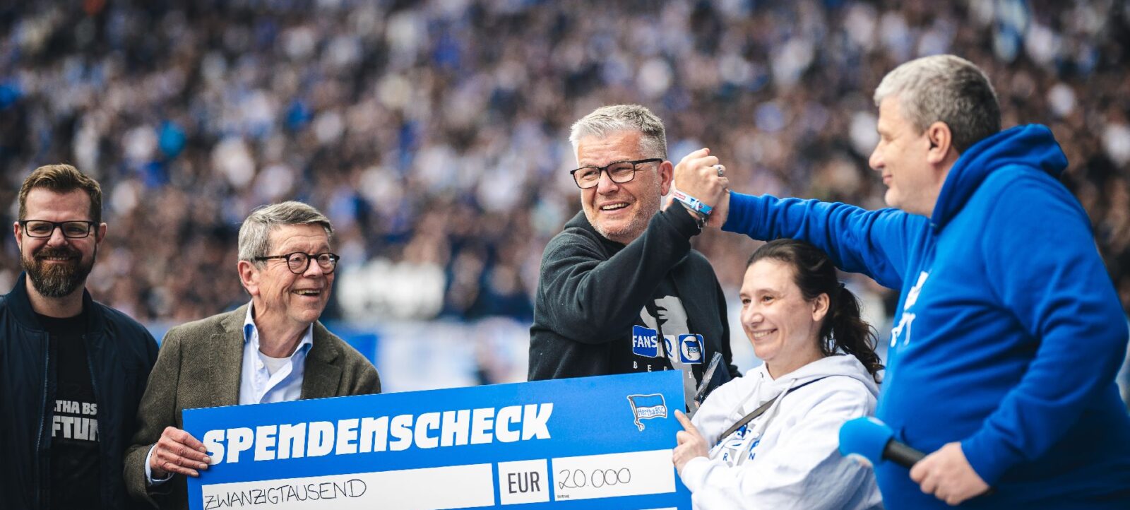 Gruppe von Personen hält einen großen Scheck über 20.000 Euro von den Fans@Hertha e.V. auf einem Fußballstadion. Die Personen lächeln und feiern die Spende, im Hintergrund sind viele Zuschauer zu sehen.