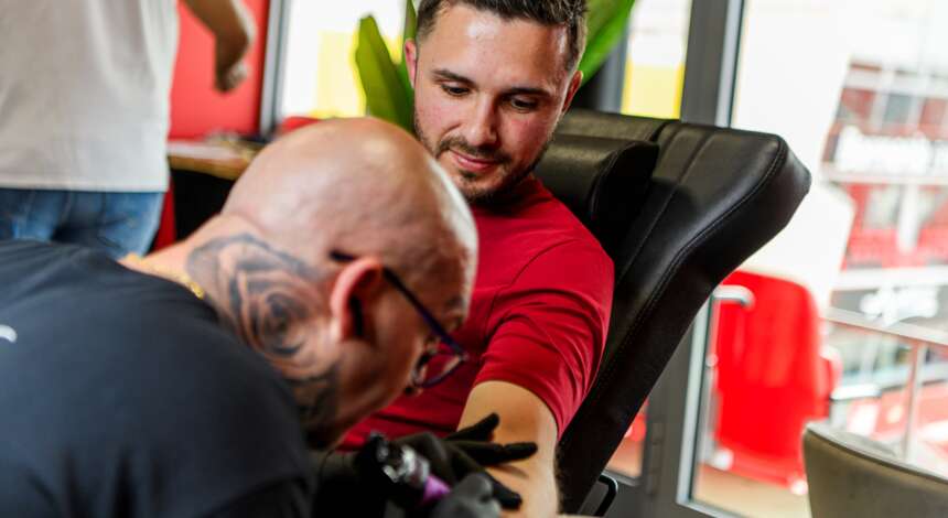 Ein Tattoo-Künstler tätowiert einen Mann, der auf einem schwarzen Ledersessel sitzt. Der Künstler trägt eine schwarze Jacke und Handschuhe, während der Mann ein rotes T-Shirt trägt. Beide scheinen sich auf den Tattoo-Prozess zu konzentrieren.