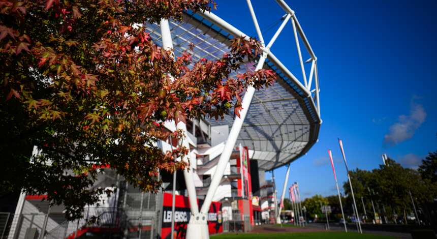 Ein modernes Fußballstadion mit weißer Stahlkonstruktion und Glasfassaden, umgeben von Bäumen mit herbstlich gefärbten Blättern. Die Sonne scheint bei klarem Himmel. Im Vordergrund sind Fahnenmasten und ein Schild mit dem Logo von Bayer 04 Leverkusen zu sehen.