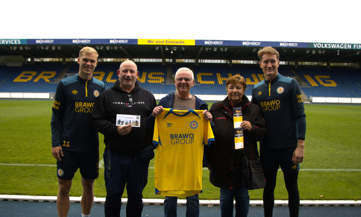 Fünf Personen stehen auf einem Fußballfeld, darunter zwei Spieler in Trainingskleidung mit dem Aufdruck "BRAWG GROUP". Zwei der Personen halten ein gelbes Trikot mit demselben Aufdruck. Im Hintergrund ist die Tribüne des Stadions mit der Aufschrift "BRAUN SCHWEIG" zu sehen. Die Gruppe scheint sich für ein gemeinsames Foto aufgestellt zu haben, möglicherweise im Rahmen eines Events oder einer Aktion.