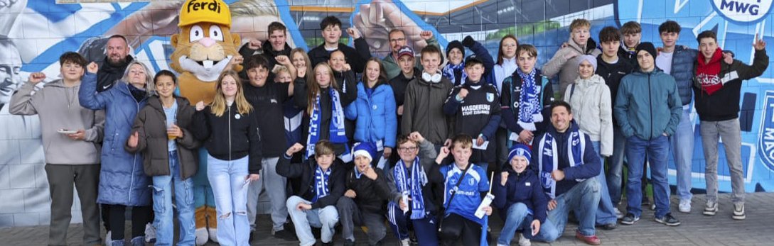 Eine Gruppe von Kindern und Erwachsenen posiert für ein Gruppenfoto vor einem blau-weißen Hintergrund mit einem Maskottchen in der Mitte. Die Kinder tragen verschiedene Kleidungsstücke, darunter Jacken, Schals und Mützen in den Vereinsfarben. Einige halten Schilder oder machen peace-Zeichen.