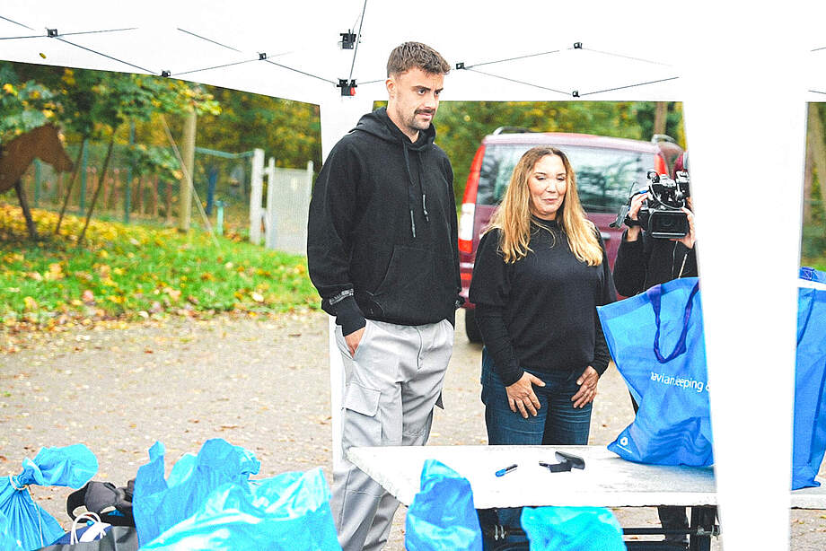 Zwei Personen stehen vor einem Pavillon im Freien, möglicherweise bei einem Event oder einer Aktion. Eine Person hält eine blaue Tüte mit Aufdruck, während eine andere Person eine Kamera bedient. Im Hintergrund sind ein rotes Auto und ein Pferd auf einer Weide zu sehen.