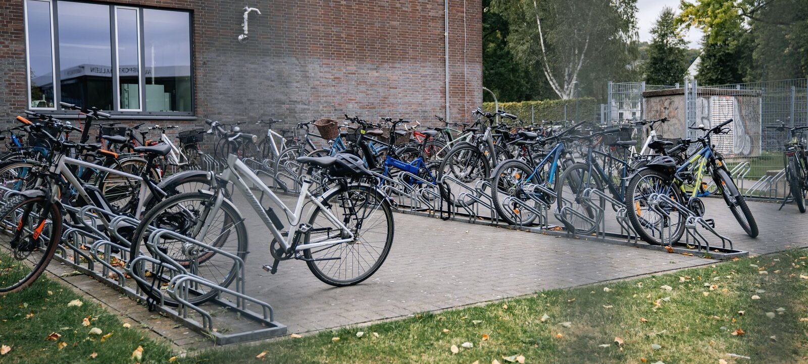 Ein überfüllter Fahrradständer vor einem Gebäude mit mehreren Fahrrädern, die an den Ständern befestigt sind. Im Hintergrund ist ein Zaun und einige Bäume zu sehen.