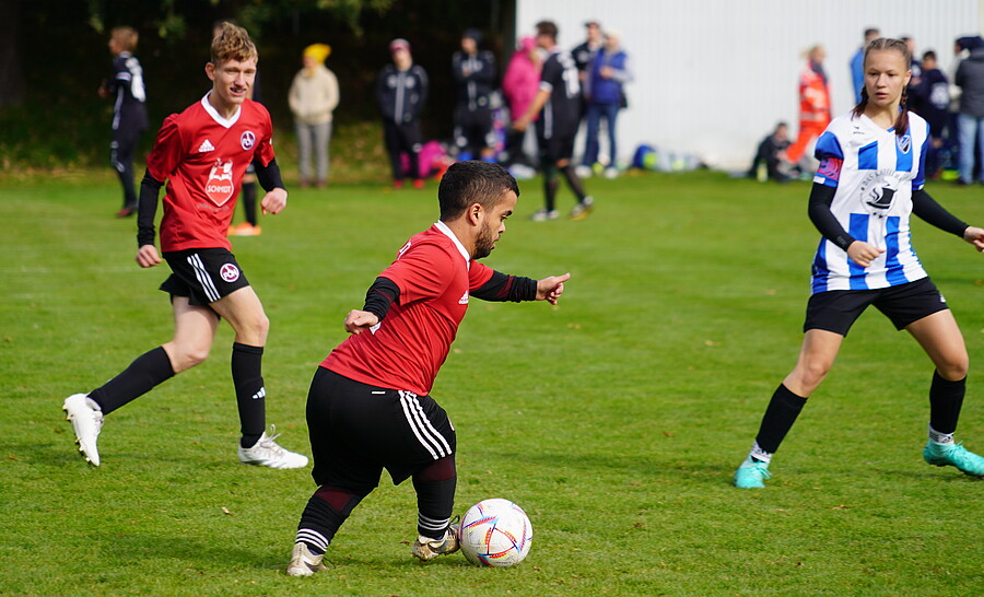 Drei Fußballspieler in Aktion auf dem Rasen. Ein Spieler in rotem Trikot kontrolliert den Ball, während zwei Gegenspieler in rotem und blau-weiß gestreiftem Trikot versuchen, den Ball zu erobern. Im Hintergrund sind weitere Personen zu sehen, die das Spiel beobachten.