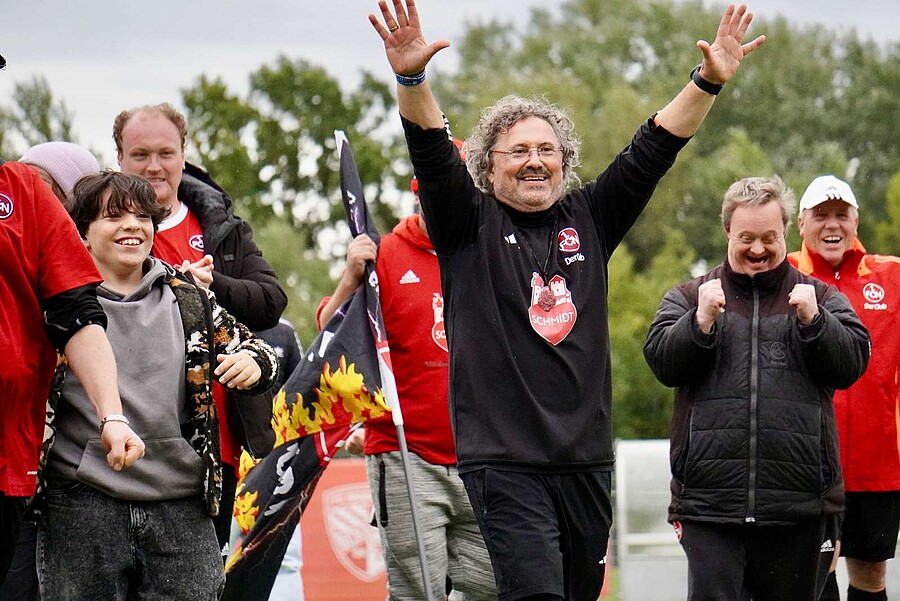 Eine Gruppe von Menschen, darunter ein Mann mit einem "Herzschild"-Logo auf seinem schwarzen T-Shirt, steht auf einem Sportplatz. Sie lächeln und winken in die Kamera. Im Hintergrund sind Bäume und ein Zaun zu sehen. Einige der Personen tragen rote Jacken mit einem Logo, das an ein Herz erinnert.