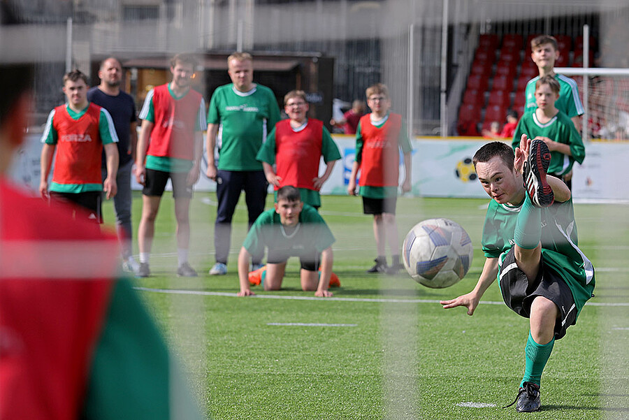 Ein junger Fußballspieler in grünem Trikot und schwarzer Hose schießt einen Fußball auf dem Rasen. Im Hintergrund stehen und knien weitere Jugendliche in roten und grünen Trikots und beobachten das Spiel.