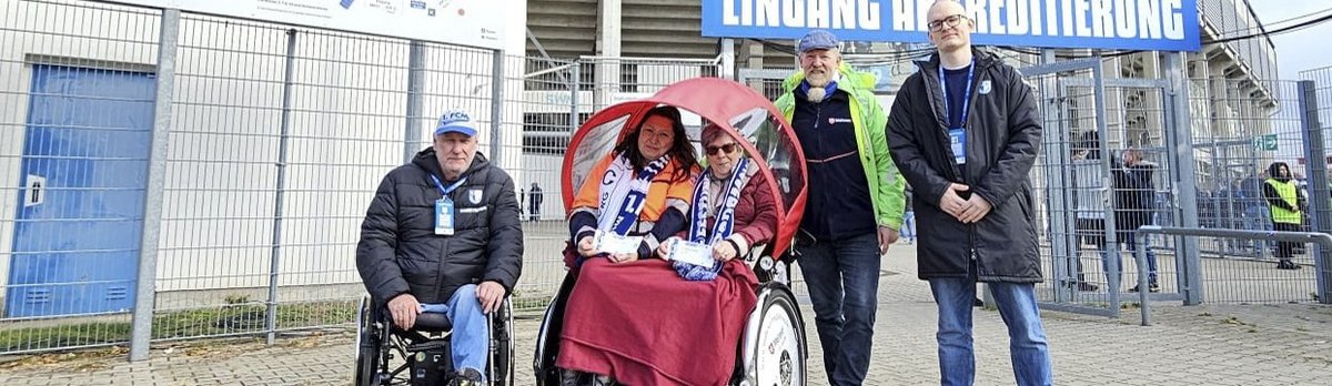 Eine Gruppe von Menschen vor einem Stadion. Eine Person im Rollstuhl wird von einer anderen Person geschoben, die einen Regenschirm hält. Zwei weitere Personen stehen daneben, eine davon trägt eine Weste. Im Hintergrund ist ein Eingang mit der Aufschrift 'Lingang Hauptredition' zu sehen.