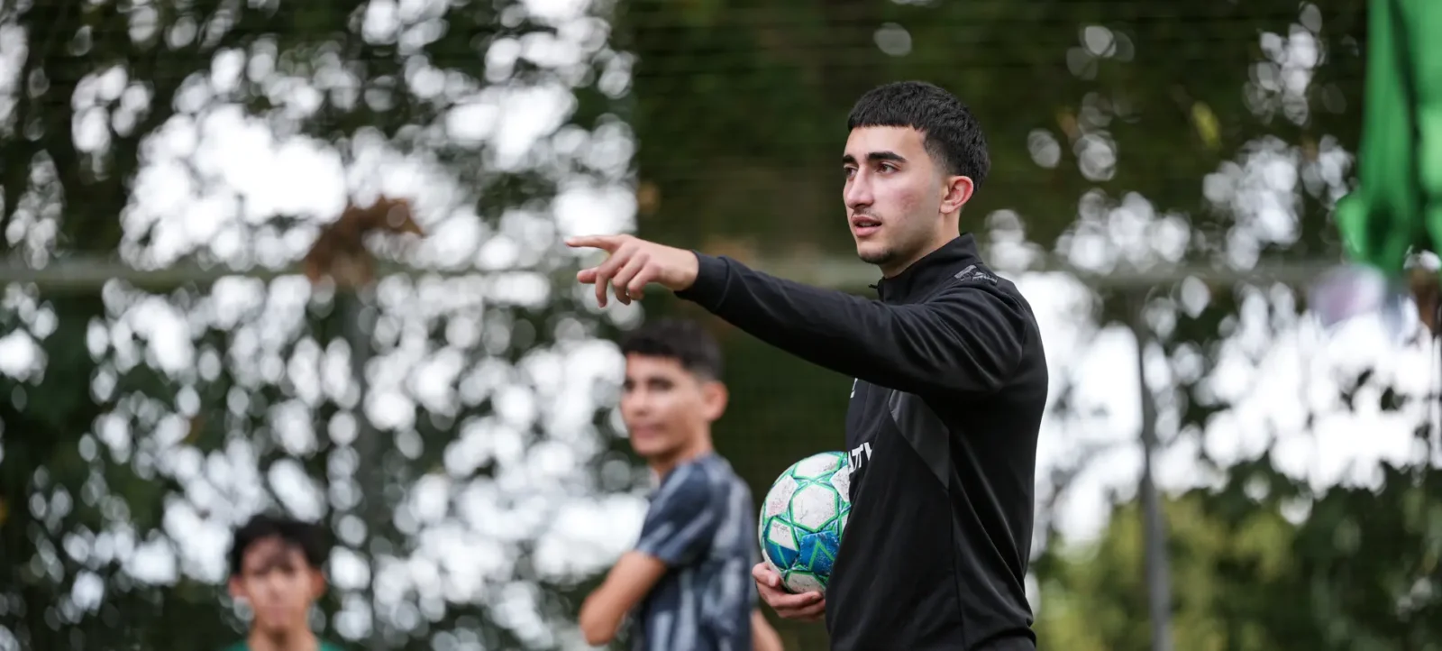Ein Trainer in schwarzer Sportkleidung hält einen Fußball in der Hand und gibt Anweisungen an junge Fußballspieler auf einem Feld. Im Hintergrund sind blühende Bäume und ein grünes Netz zu sehen.
