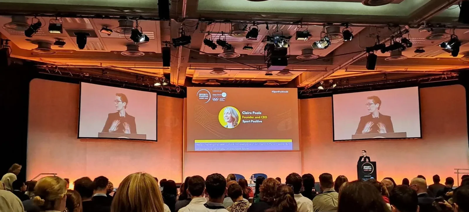 Eine Person steht auf einer Bühne und hält eine Präsentation in einem gut beleuchteten Saal. Die Person wird auf zwei großen Bildschirmen gezeigt, die an den Seiten der Bühne angebracht sind. Der Name und die Position der Person, Claire Poole, Gründerin und CEO von Sport Positive, sind auf den Bildschirmen angezeigt. Das Publikum sitzt auf Stühlen und blickt zur Bühne.