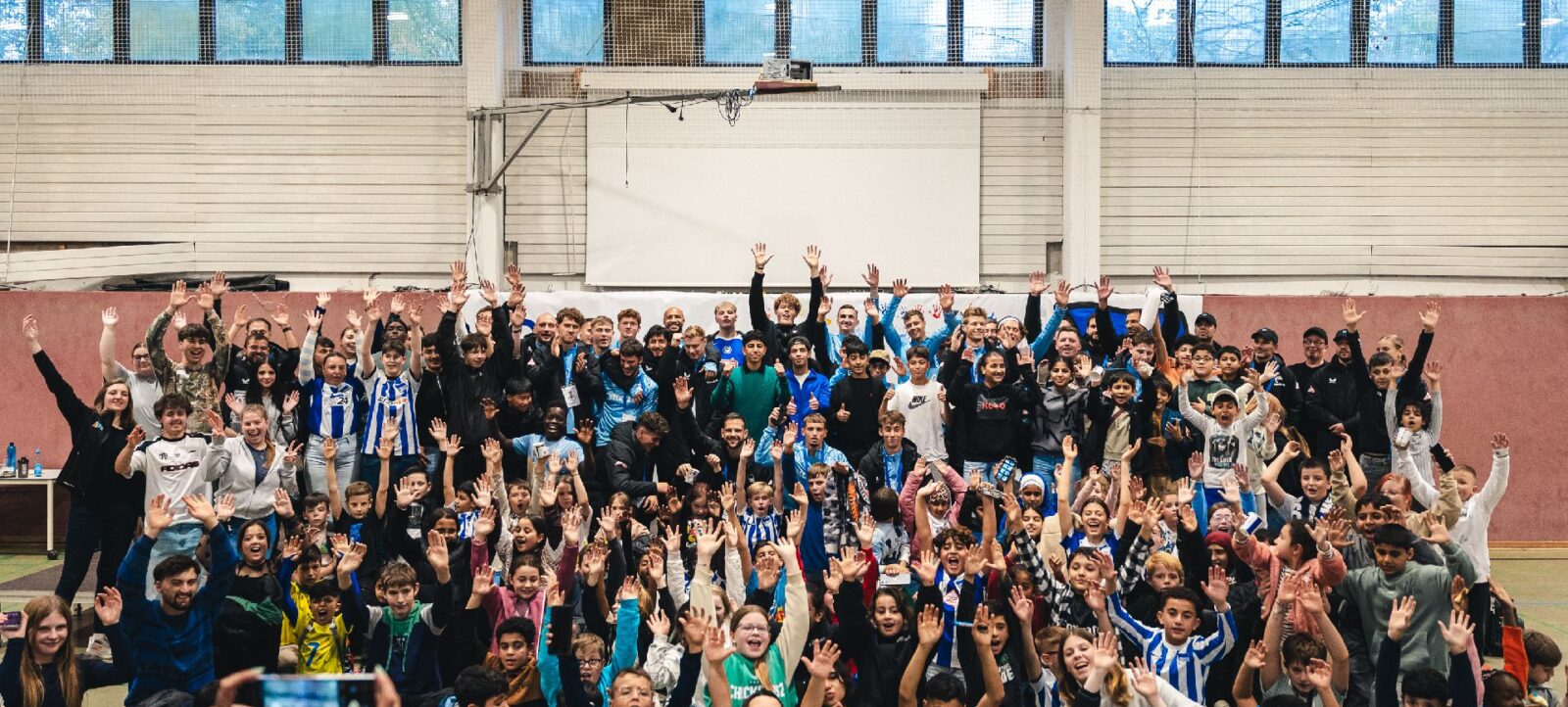 Eine große Gruppe von Kindern und Betreuern in einer Sporthalle, alle lächeln und winken freudig in die Kamera. Die Kinder tragen verschiedene Sport- und Freizeitkleidung, darunter auch T-Shirts mit Vereinslogos. Die Atmosphäre ist fröhlich und gesellig, was auf ein gemeinsames Erlebnis oder eine Veranstaltung hindeutet.