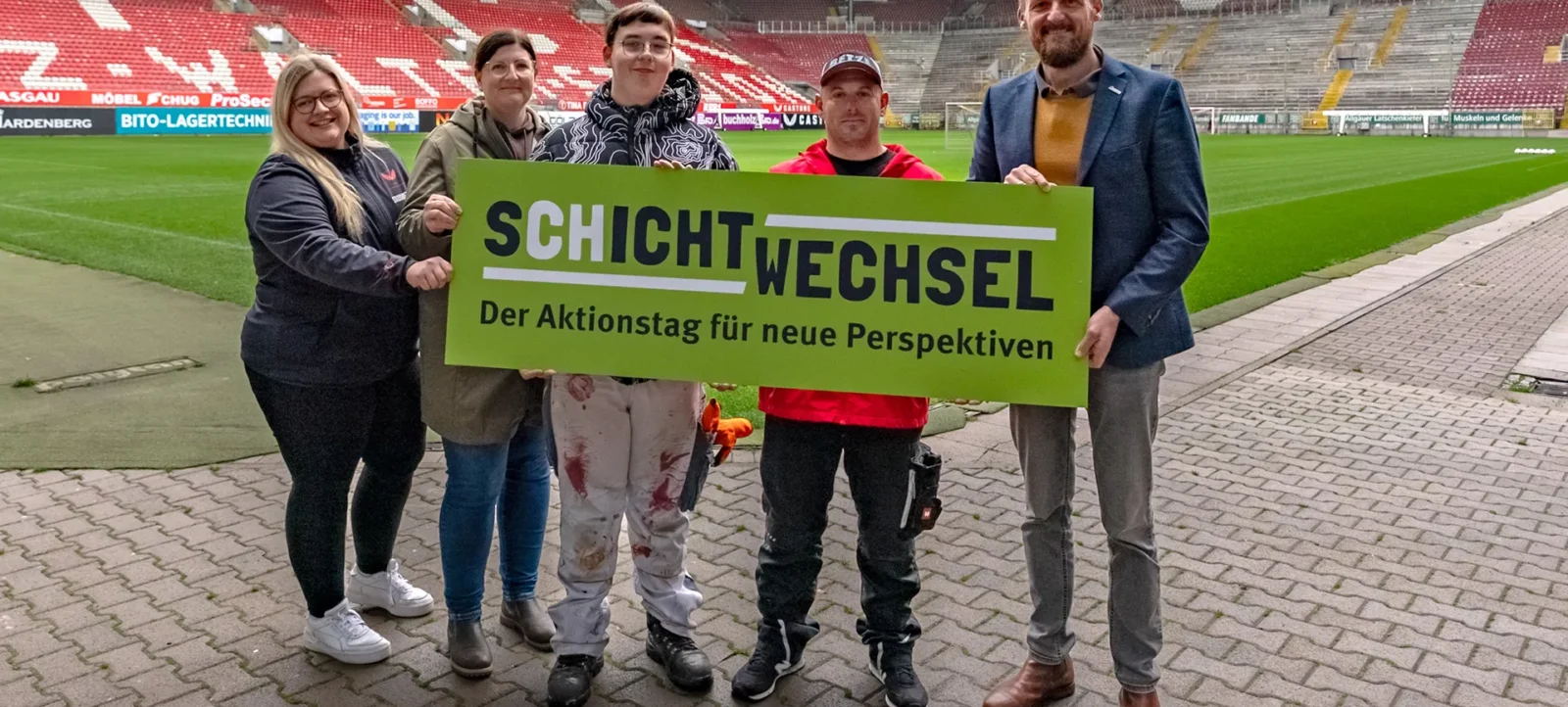 Fünf Personen stehen auf einem Fußballplatz und halten ein grünes Schild mit der Aufschrift "SCHICHTWECHSEL - Der Aktionstag für neue Perspektiven". Im Hintergrund sind die Tribünen des Stadions zu sehen.