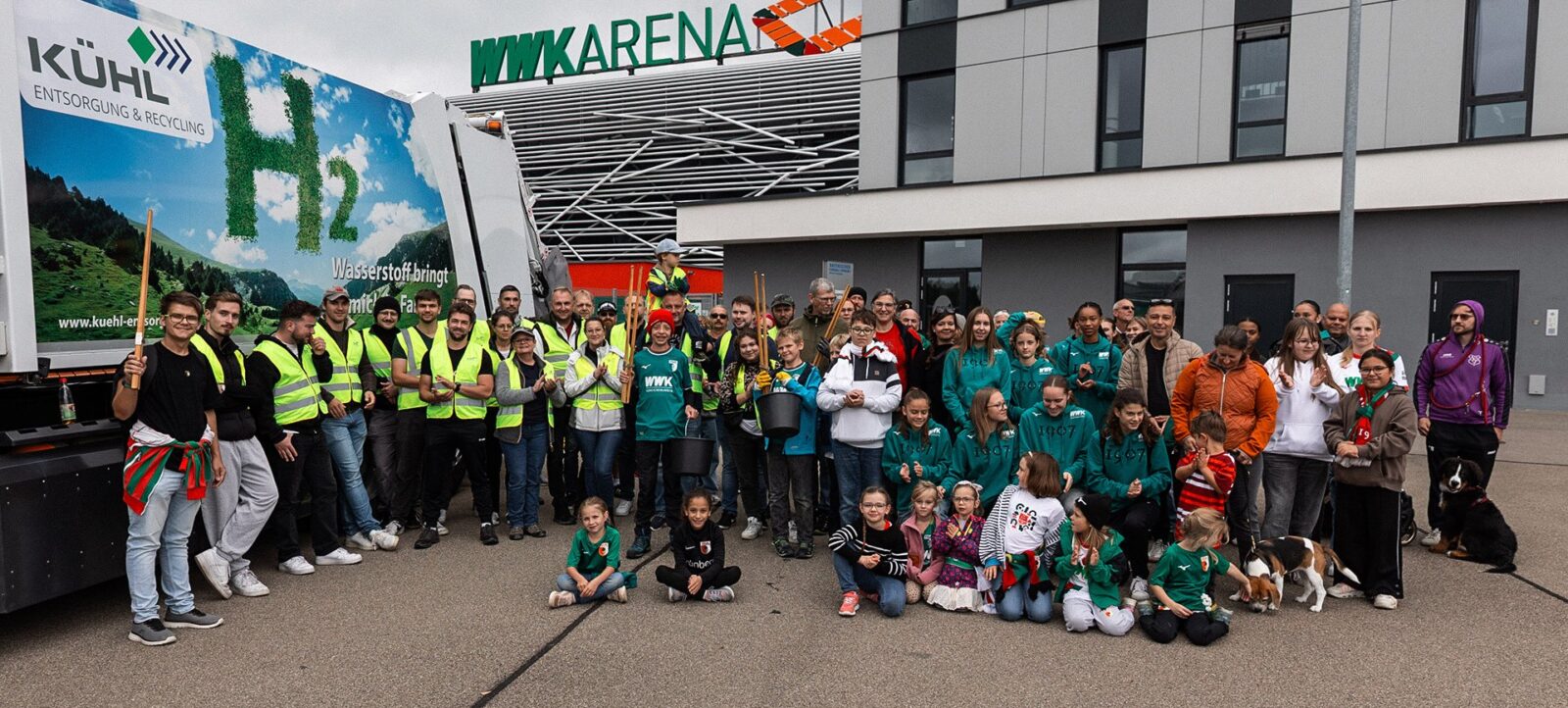 Eine Gruppe von Menschen, darunter Erwachsene und Kinder, steht vor der WWK Arena. Sie tragen gelbe Sicherheitswesten und T-Shirts mit dem Aufdruck "WWK". Einige halten Besen und Schaufeln in den Händen. Im Hintergrund ist ein Plakat mit der Aufschrift "Kühl Entsorgung & Recycling" und einem Hinweis auf Wasserstoff zu sehen. Die Atmosphäre wirkt fröhlich und engagiert, möglicherweise handelt es sich um eine Gemeinschaftsaktion oder ein Event im Rahmen eines Nachhaltigkeitsprojekts.