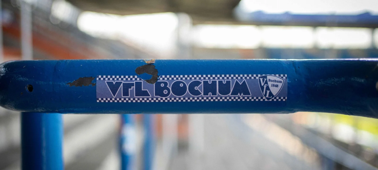 Ein blaues Metallrohr mit einem Aufkleber des Vereins VfL Bochum. Der Aufkleber zeigt das Vereinslogo und den Vereinsnamen. Das Rohr ist Teil einer Struktur, möglicherweise eines Spielplatzgeräts oder einer Tribüne.