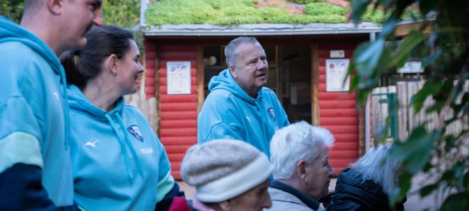 Drei Personen in blauen Jacken stehen vor einem roten Holzgebäude und unterhalten sich mit drei älteren Menschen, die auf einer Bank sitzen. Die Szene spielt im Freien, umgeben von Grünflächen und Bäumen.