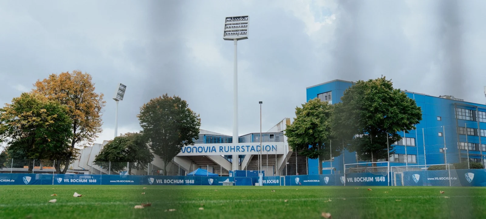 Ein Fußballfeld mit Blick auf das Stadion "Vonovia Ruhrstadion". Im Vordergrund ist der Rasen mit einigen Blättern bedeckt, und im Hintergrund sind die Tribünen und ein blaues Gebäude zu sehen. Zwei große Flutlichtmasten ragen in den Himmel.