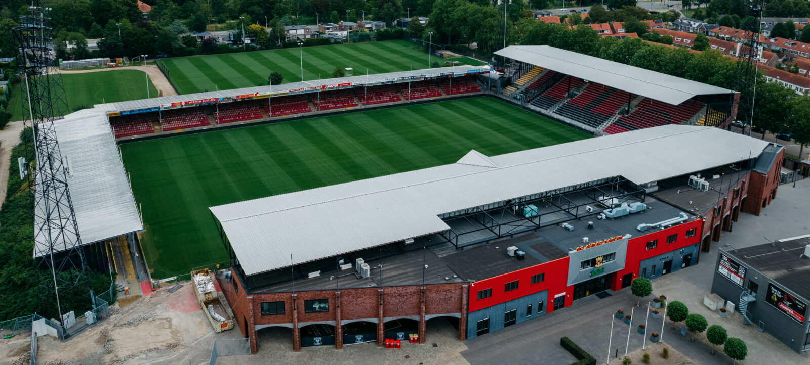Luftaufnahme eines Fußballstadions mit grünem Rasen und roten Sitzplätzen. Das Stadion ist von einer Wohngegend und viel Grün umgeben. Die Haupttribüne hat ein graues Dach und ist mit Werbebannern verziert. Im Vordergrund sind Gebäude und ein Zaun zu sehen.