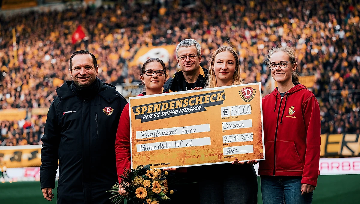 Eine Gruppe von vier Personen hält einen großen Scheck über 5.000 Euro von der SG Dynamo Dresden für den Verein Moosmützel-Hof e.V. Sie stehen auf einem Fußballfeld vor einer großen Zuschauermenge und lächeln in die Kamera. Eine Person hält einen Blumenstrauß.