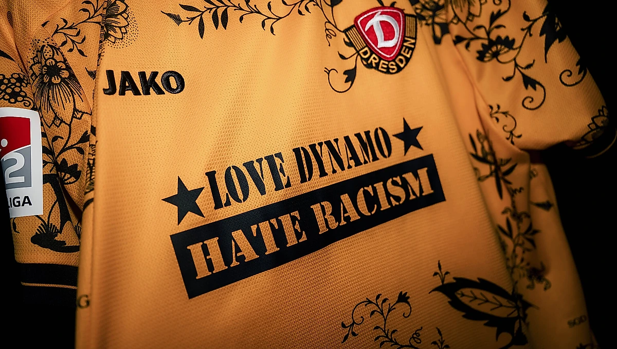 Ein Trikot mit dem Aufdruck "Love Dynamo, Hate Racism" und dem Logo des Vereins 1. FC Nürnberg. Das Trikot hat ein goldbraunes Design mit schwarzen floralen Mustern und dem Sponsorenlogo "JAKO".