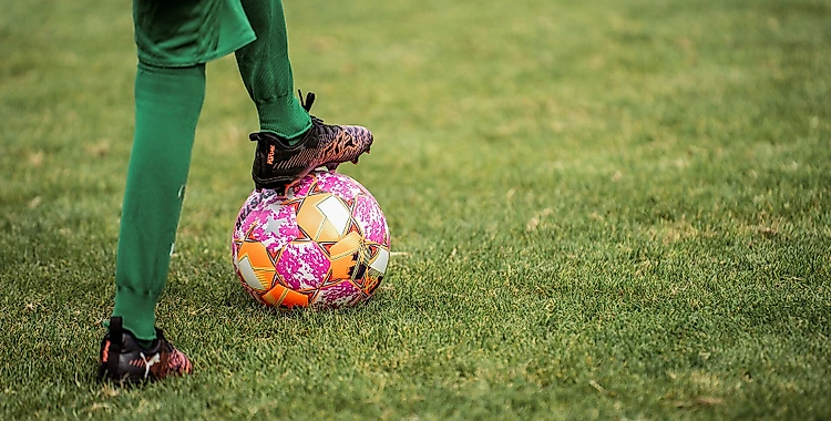 Ein Fußballspieler in grüner Hose und schwarzen Fußballschuhen steht auf einem Rasenplatz und berührt mit dem Fuß einen bunten Fußball.