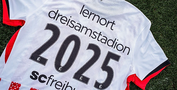 Ein weißes Trikot mit roten Ärmeln liegt auf einem Rasenboden. Auf dem Rücken des Trikots steht in großen schwarzen Zahlen die Zahl "2025". Darüber ist in kleinerer Schrift "lernort dreisamstadion" und darunter "sc freiburg" zu lesen.