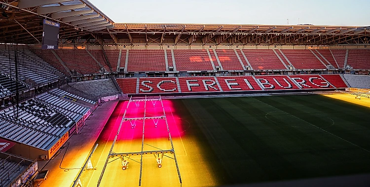 Ein leeres Fußballstadion mit roten Sitzplätzen und dem Schriftzug "SC Freiburg" auf der Gegengerade. Das Spielfeld ist beleuchtet, wobei ein Teil des Rasens in gelbes und pinkes Licht getaucht ist.