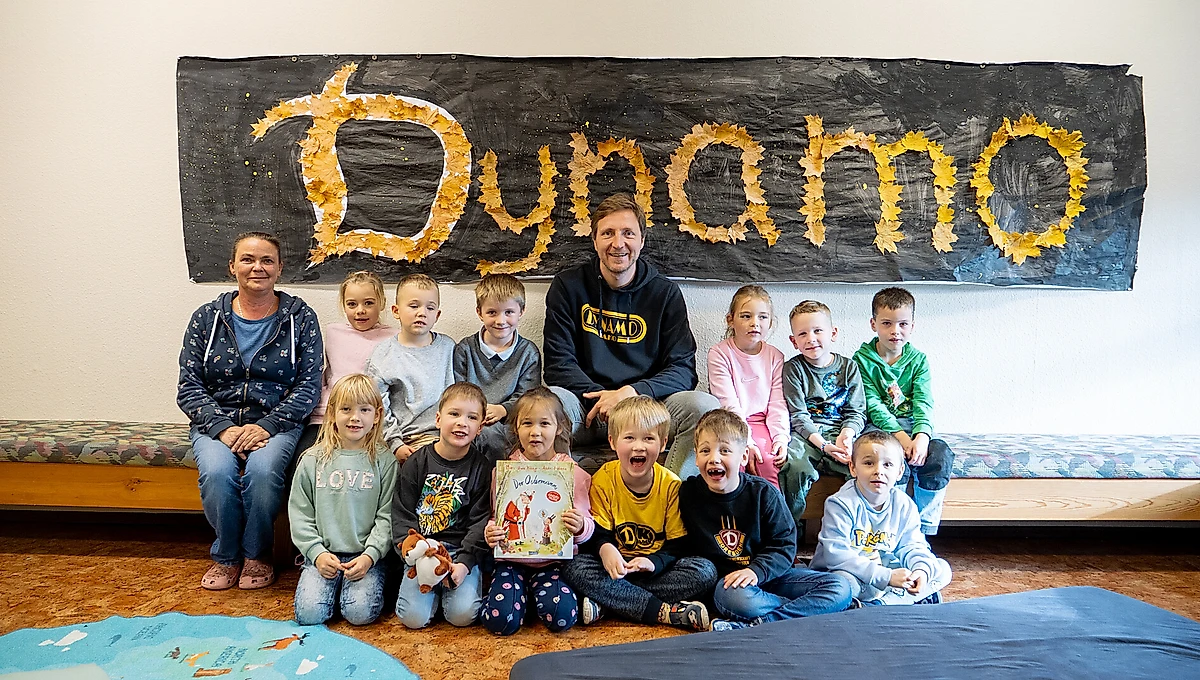 Eine Gruppe von Kindern und zwei Erwachsenen sitzen vor einem selbstgemachten Schild mit der Aufschrift "Dunno" aus Blättern. Die Kinder halten ein Buch und ein Stofftier. Die Erwachsenen und einige Kinder tragen Kleidung mit dem Aufdruck "DMD". Die Szene findet in einem Raum mit bunten Teppichen statt.