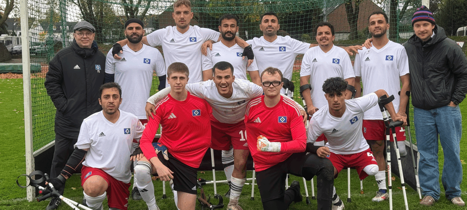 Eine Gruppe von Menschen mit Behinderungen, speziell Amputierten-Fußball-Spielern, posieren vor einem Tor. Sie tragen Trikots mit dem Logo des Hamburger SV und lächeln in die Kamera. Die Spieler sind sowohl in weißen als auch in roten Trikots gekleidet und einige von ihnen verwenden Prothesen. Zwei Begleitpersonen stehen links und rechts neben der Gruppe. Das Foto wurde auf einem Fußballfeld aufgenommen.
