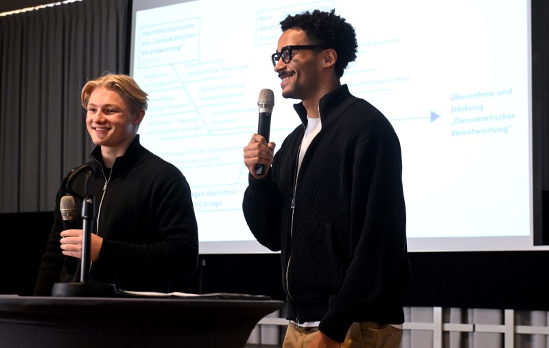 Zwei Personen stehen an einem Pult und halten eine Präsentation zum Thema Antirassismus. Eine Person spricht in ein Mikrofon, während die andere lächelnd zuhört. Im Hintergrund ist eine Projektionsfläche mit Text zu sehen.