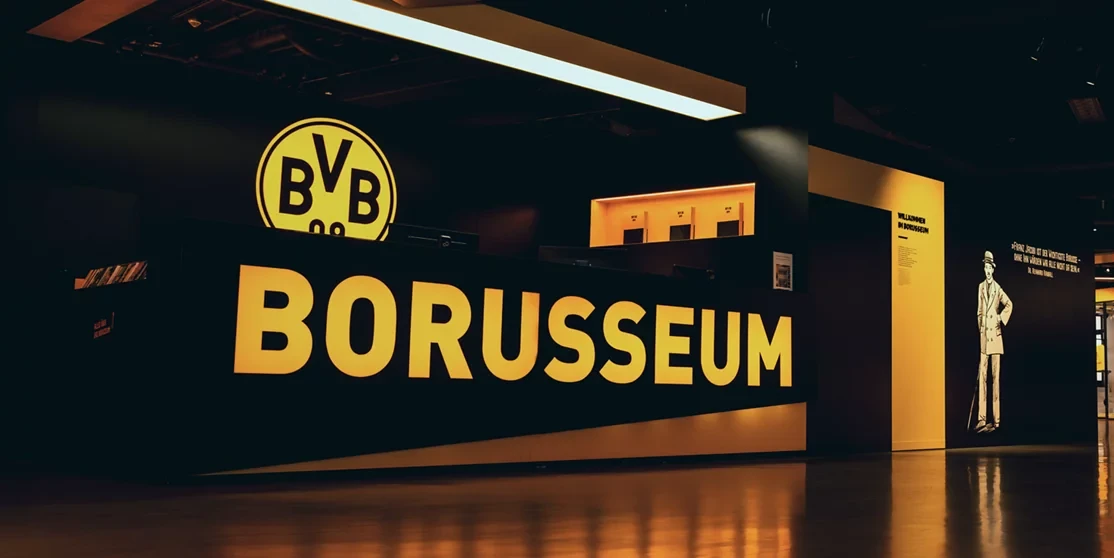 Eingangsbereich des Borusseums mit dem großen Logo des BVB und der Aufschrift 'BORUSSEUM' in gelben Buchstaben auf schwarzem Hintergrund. Rechts im Bild ist eine Silhouette einer Person mit Hut zu sehen.