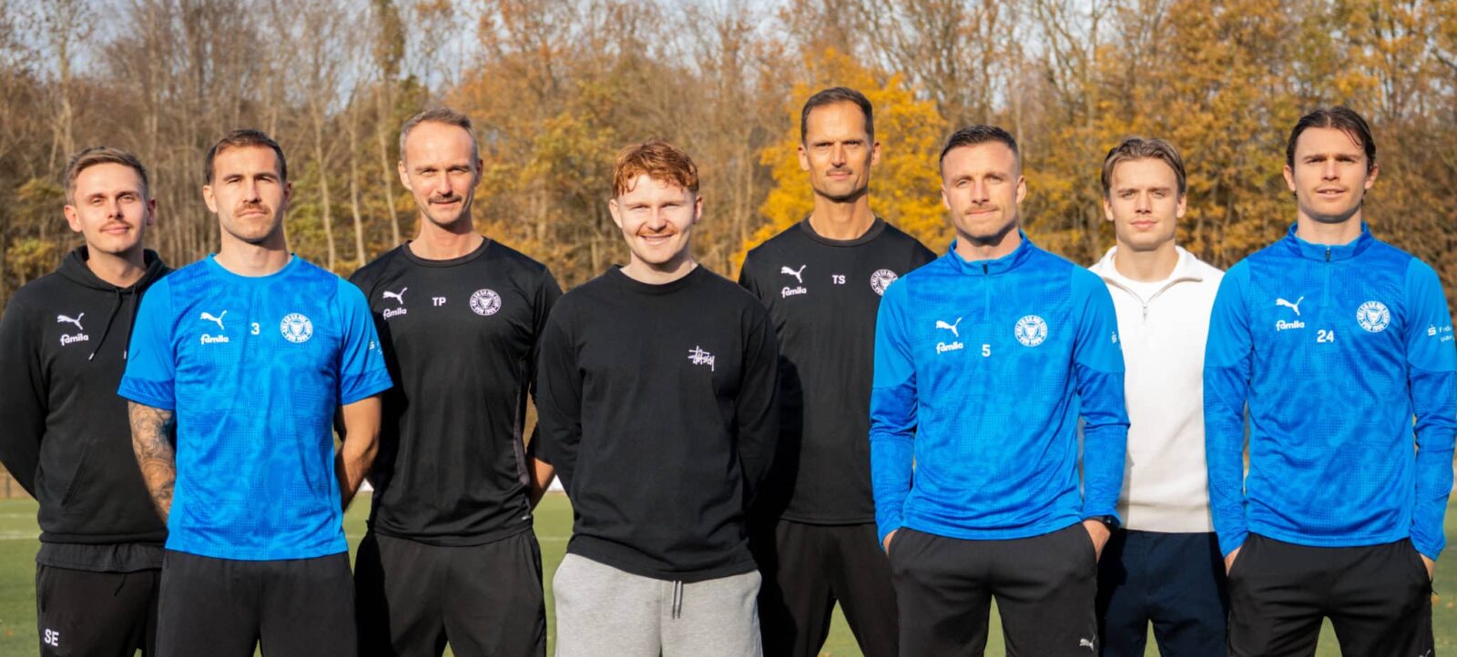 Eine Gruppe von sieben Personen, darunter Spieler und Trainer, posieren für ein Gruppenfoto auf einem Fußballplatz. Die Spieler tragen blaue Trainingsjacken mit dem Logo von Eintracht Braunschweig, während die Trainer schwarze Jacken mit dem gleichen Logo tragen. Im Hintergrund sind Bäume mit herbstlichem Laub zu sehen.