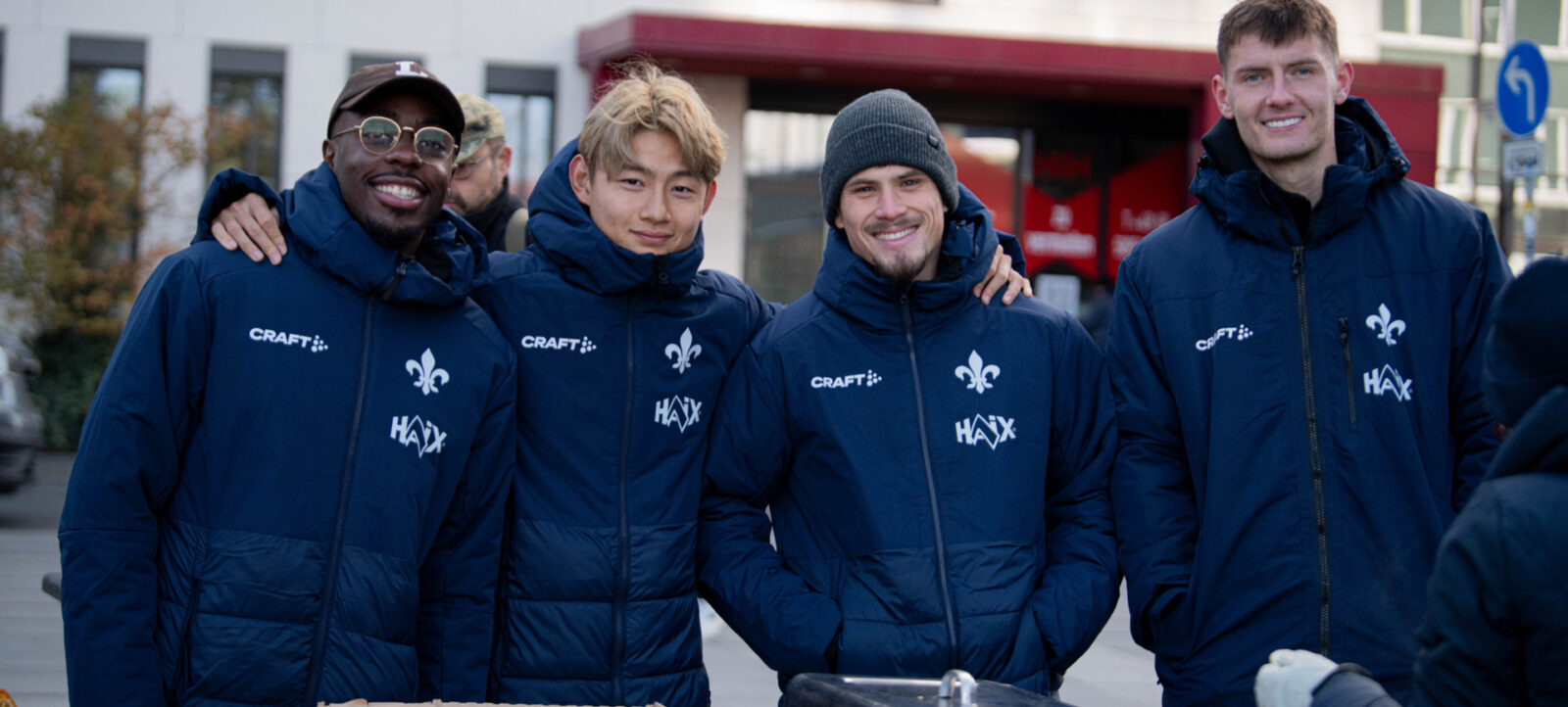 Vier Personen in blauen Jacken mit dem Logo "HAIX" und einem Lilien-Emblem stehen lächelnd nebeneinander. Sie scheinen sich für ein Foto zu gruppieren, möglicherweise bei einem sozialen oder gemeinnützigen Event. Im Vordergrund befindet sich ein Tisch mit einer offenen Kartonschachtel und einem Metallbehälter, was auf eine Essensausgabe oder eine ähnliche Aktivität hindeutet.
