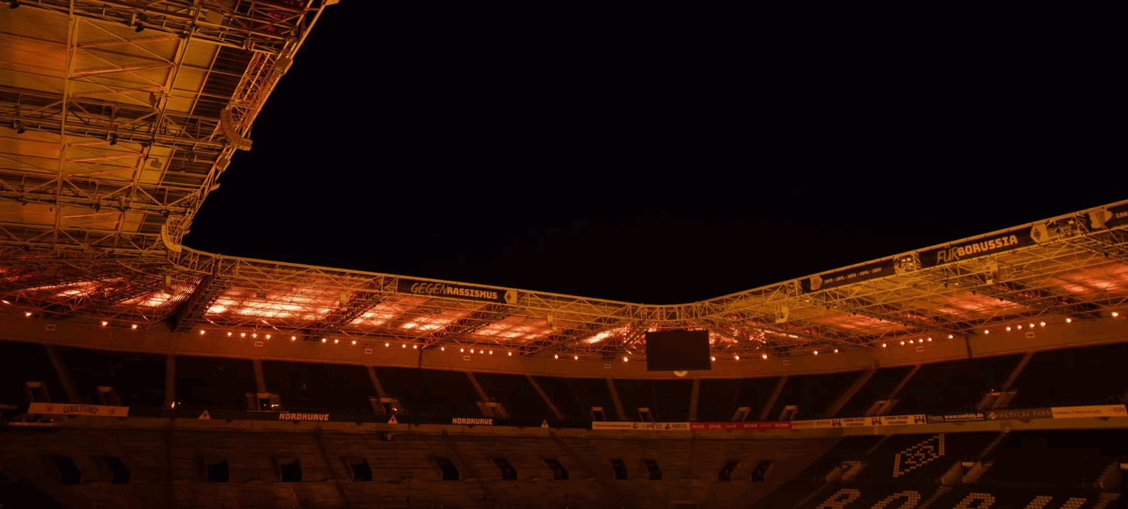 Ein leeres Fußballstadion bei Nacht, beleuchtet von gelbem Licht. Die Tribünen sind dunkel und leer, und ein Banner mit der Aufschrift "Gegen Rassismus" ist sichtbar. Die Struktur des Stadions ist deutlich zu erkennen, mit Metallträgern und Sitzreihen.