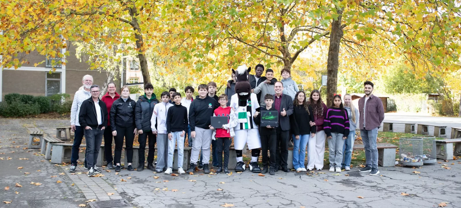 Eine Gruppe von Menschen, darunter Kinder und Erwachsene, steht im Freien vor einem Hintergrund aus Bäumen mit herbstlich gefärbten Blättern. In der Mitte der Gruppe befindet sich eine Person in einem Maskottchen-Kostüm, das an ein Pferd erinnert. Einige der Kinder halten Laptops in den Händen und zeigen ihre Freude mit einem Daumen-hoch-Zeichen. Die Gruppe scheint Teil eines Gemeinschafts- oder Bildungsprojekts zu sein, möglicherweise im Zusammenhang mit Nachhaltigkeit oder Technologie.