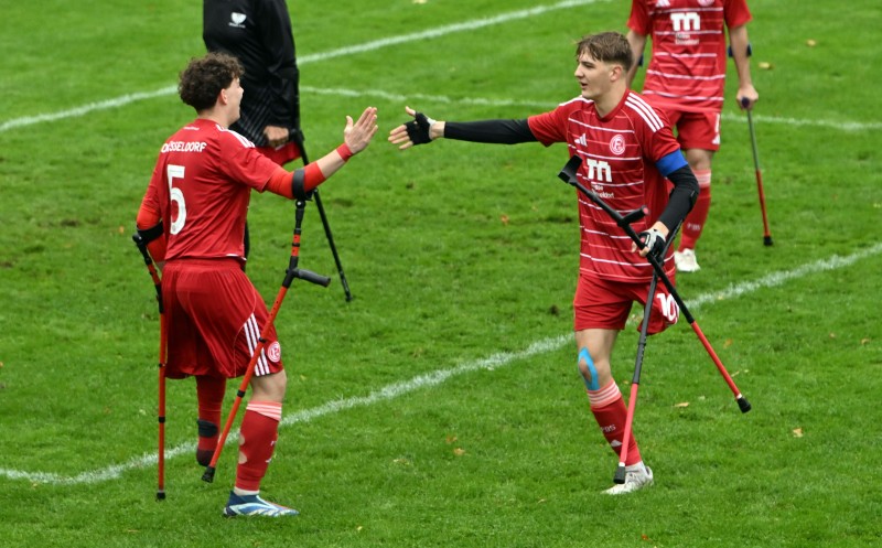 Zwei Fußballspieler in roten Trikots mit Krücken auf dem Spielfeld, die sich gegenseitig abklatschen.