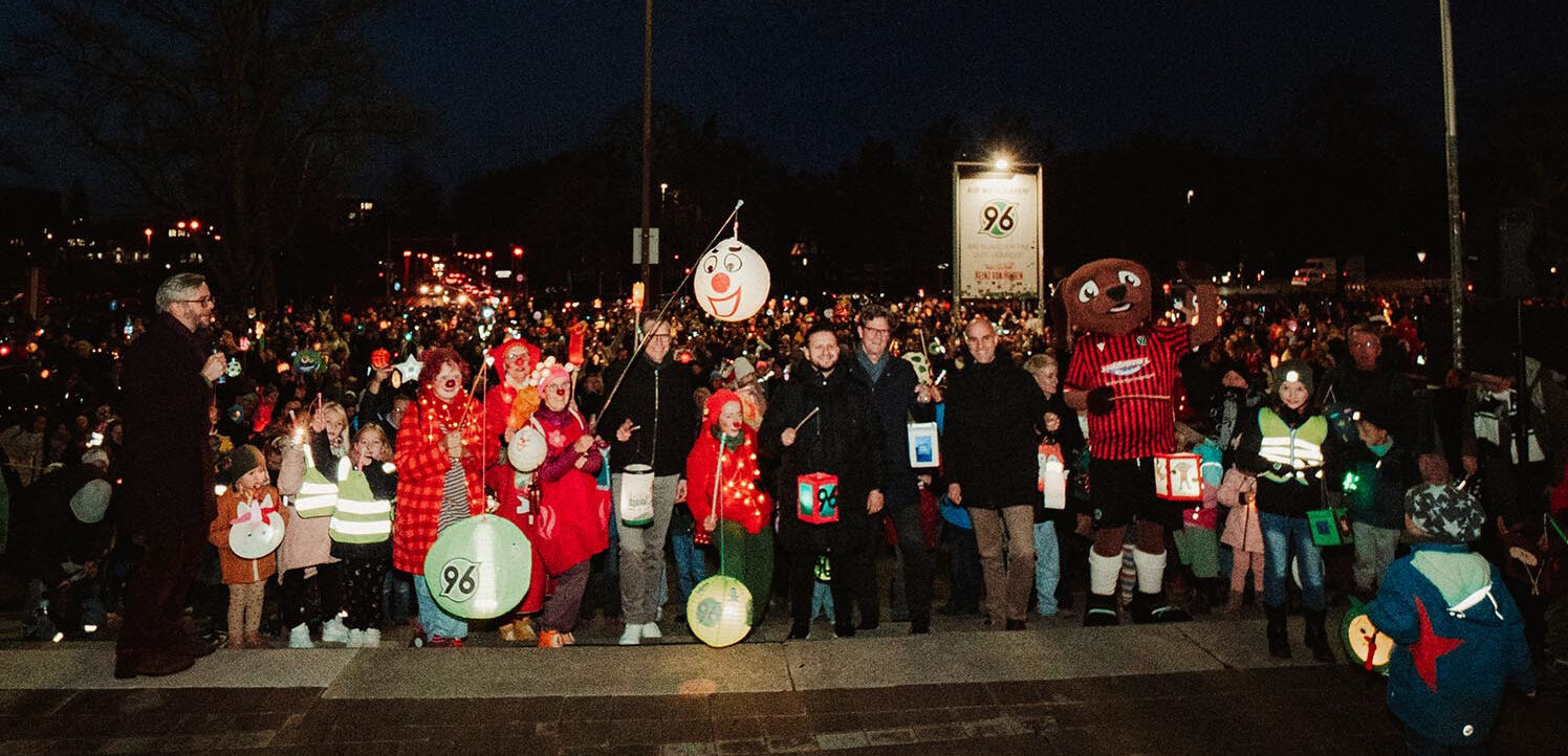 Eine große Gruppe von Menschen, darunter Kinder in Kostümen und ein Maskottchen, nehmen an einem Laternenumzug teil. Sie halten Laternen mit der Zahl 96 und bunte Luftballons. Die Veranstaltung findet nachts statt und ist gut besucht, mit vielen Teilnehmern im Hintergrund.