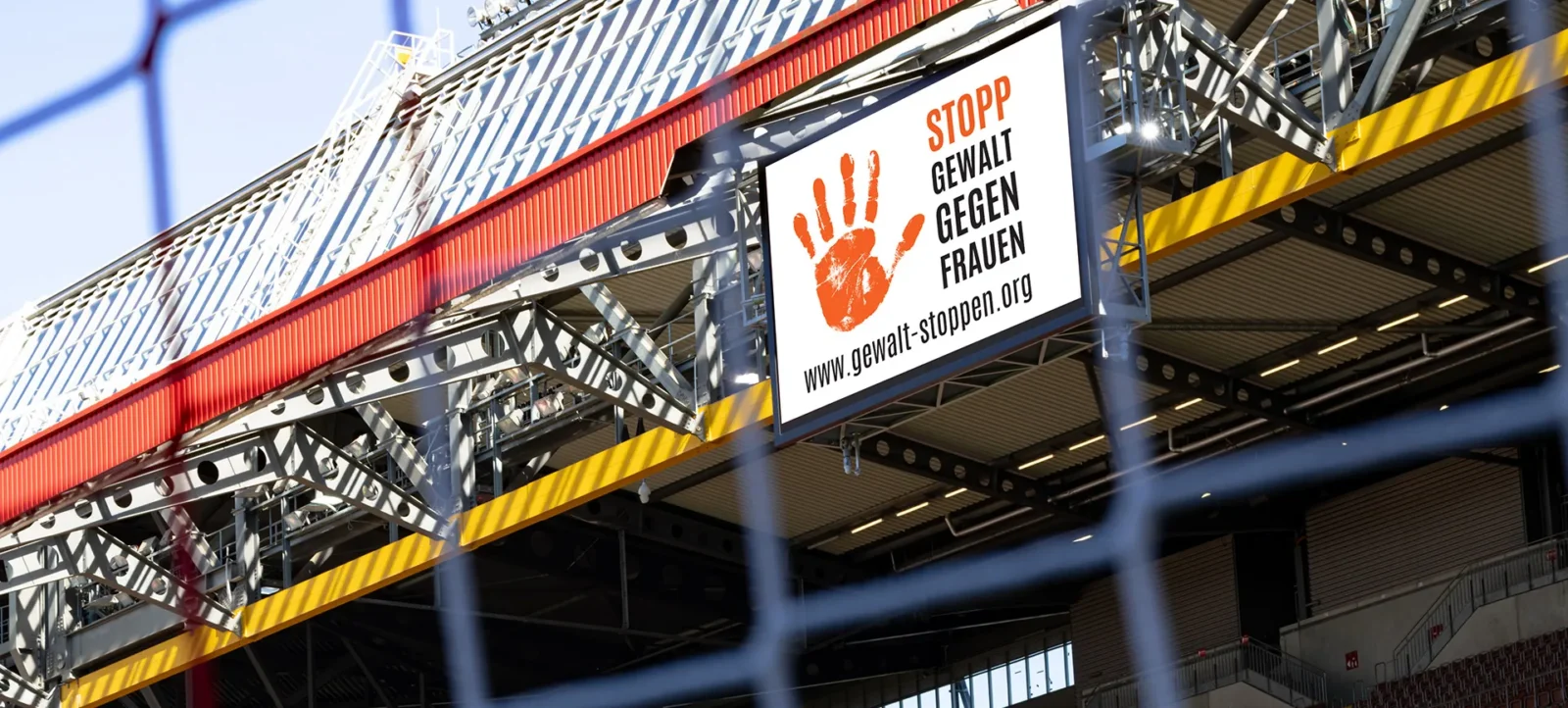 Ein Stadion mit einem Banner, das eine Kampagne gegen Gewalt an Frauen unterstützt. Das Banner zeigt eine rote Handabdruck und den Text "STOPP GEWALT GEGEN FRAUEN" sowie die Website "www.gewalt-stoppen.org".