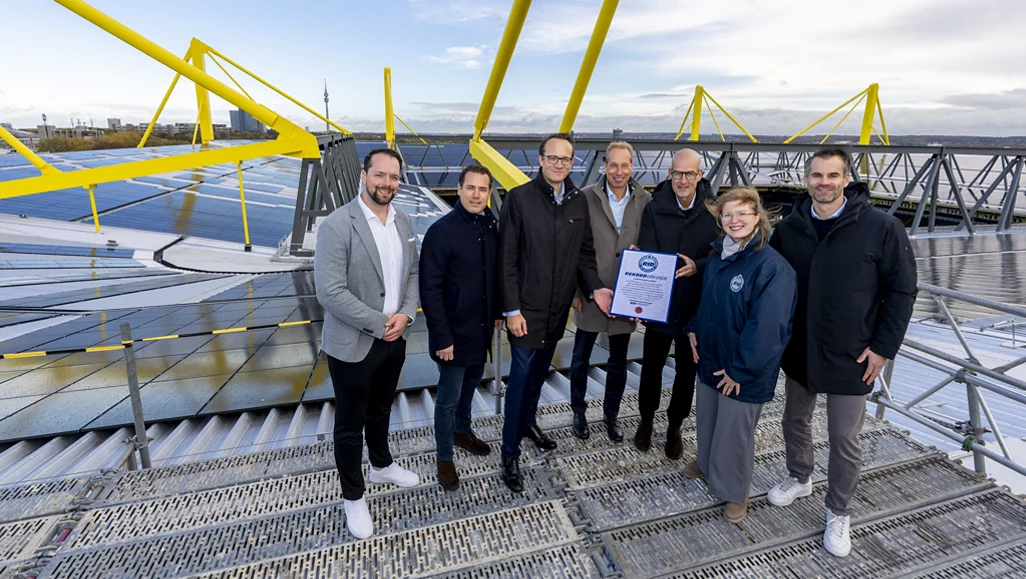 Eine Gruppe von sieben Personen steht auf einer großen Solaranlage mit gelben Stahlträgern im Hintergrund. Sie halten gemeinsam ein Zertifikat und lächeln in die Kamera. Die Solaranlage ist auf dem Dach eines Gebäudes installiert, und im Hintergrund sind Teile einer Stadt mit einem Fernsehturm zu sehen.