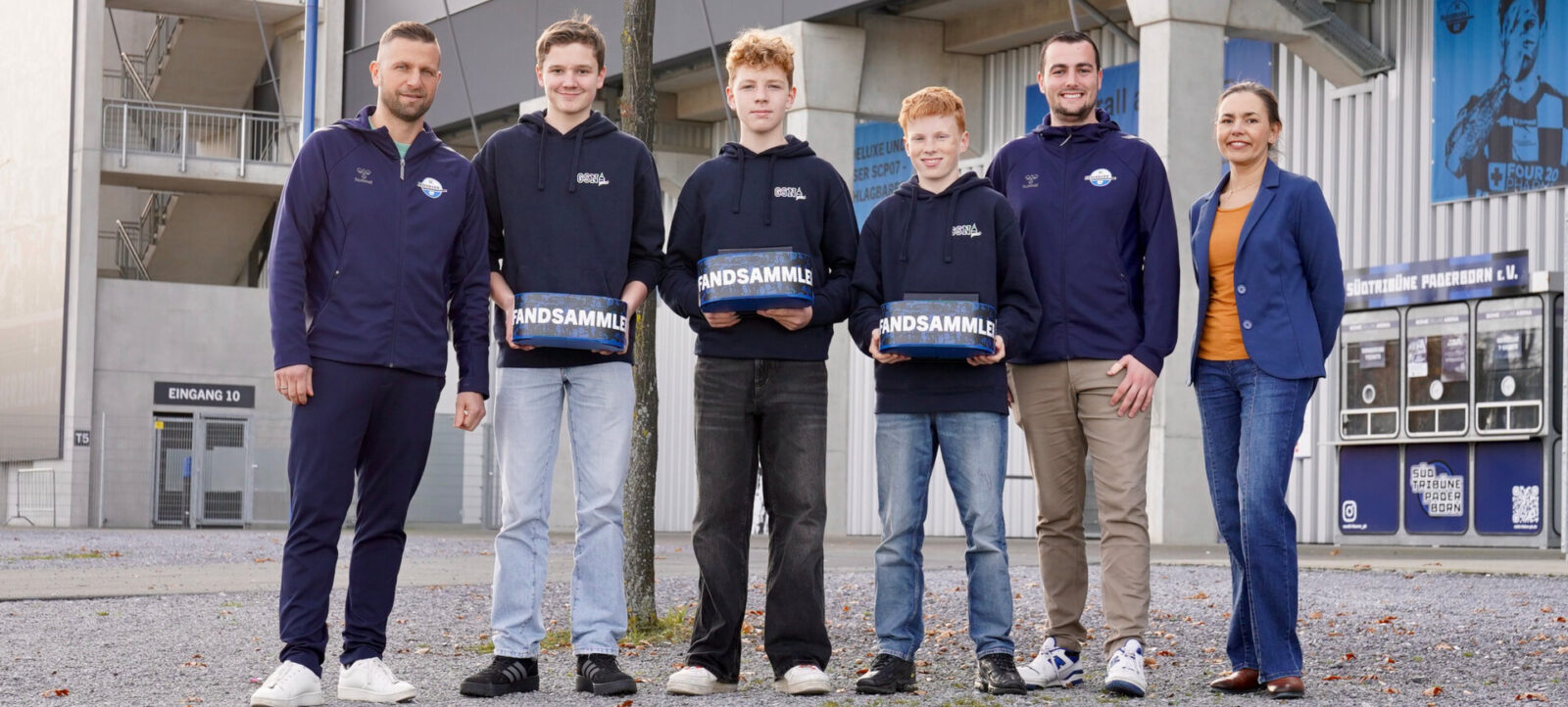 Eine Gruppe von sechs Personen, darunter vier Jugendliche und zwei Erwachsene, steht vor einem Stadion. Sie halten blaue Pfandsammelbehälter mit der Aufschrift "PFANDSAMMLER" in den Händen. Alle tragen dunkle Kleidung, einige mit Logos eines Vereins oder Projekts. Im Hintergrund ist ein Teil des Stadioneingangs mit der Bezeichnung "EINGANG 10" und Werbebanner zu sehen.