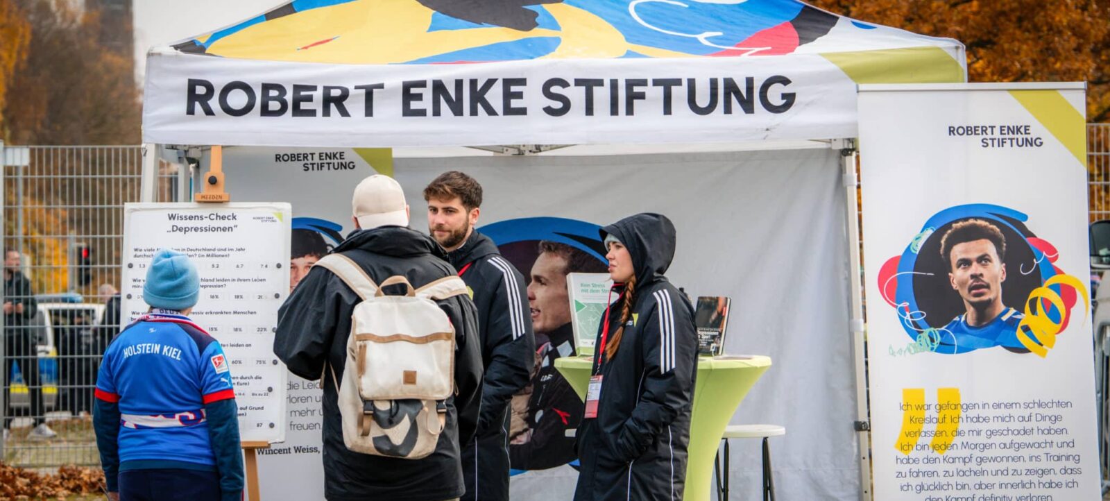 Ein Zelt der Robert Enke Stiftung mit mehreren Personen, die sich über Informationsmaterial zu Depressionen austauschen. Auf der rechten Seite des Zeltes ist ein Bild von Robert Enke mit einem Zitat von Dele Alli, einem Fußballspieler, zu sehen. Im Hintergrund trägt eine Person ein Trikot des Vereins Holstein Kiel.