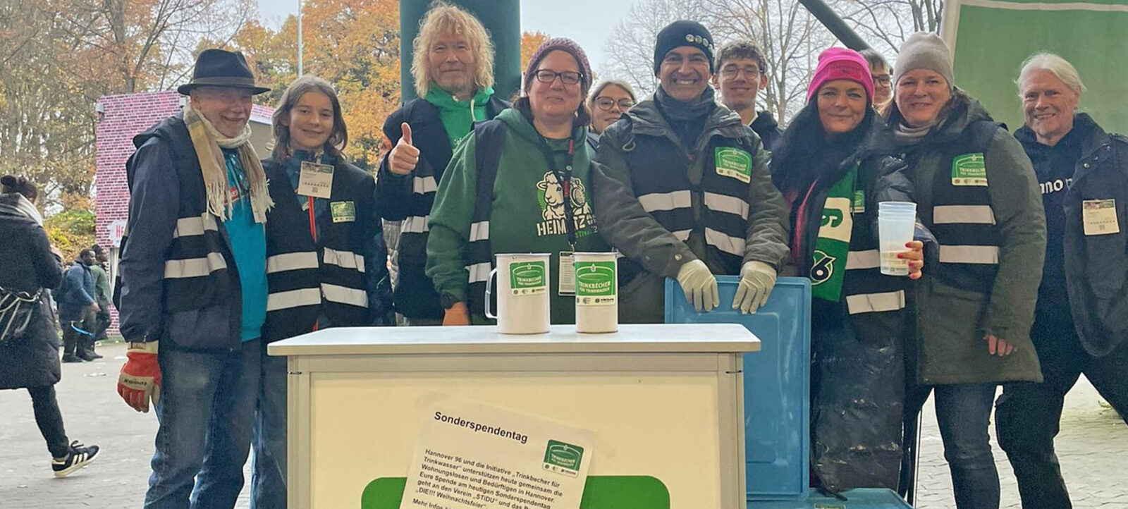 Eine Gruppe von Menschen steht vor einem Stand und lächelt in die Kamera. Sie tragen Reflektorenwesten und halten Becher in den Händen. Der Stand hat ein Schild mit der Aufschrift "Sonderspendentag" und ist mit grünen Bechern und einem Schild mit einem Logo dekoriert. Die Menschen wirken fröhlich und engagiert, möglicherweise bei einer Spendenaktion oder einem Nachhaltigkeitsprojekt.