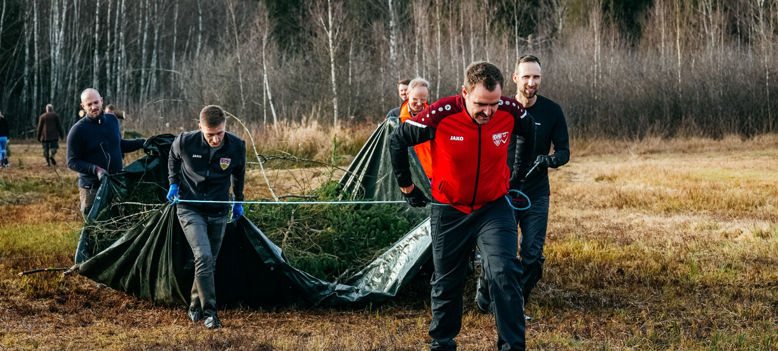 Eine Gruppe von Menschen, darunter ein Mann in einem roten Jacko-Pulli mit dem Vereinslogo des VfB Stuttgart, arbeiten gemeinsam im Freien. Sie ziehen Äste und Zweige in einen großen schwarzen Sack, um die Natur zu säubern. Die Szene spielt in einem Waldgebiet mit kahlen Bäumen und trockenem Gras, was auf eine Herbst- oder Winterzeit hindeutet.