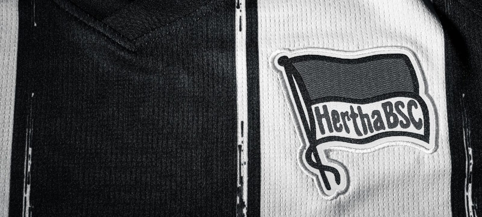 Schwarz-weiß Foto eines Trikots mit dem Logo des Hertha BSC. Das Logo zeigt ein schwarzes Banner mit der Aufschrift 'Hertha BSC' und einem großen 'S' darunter.