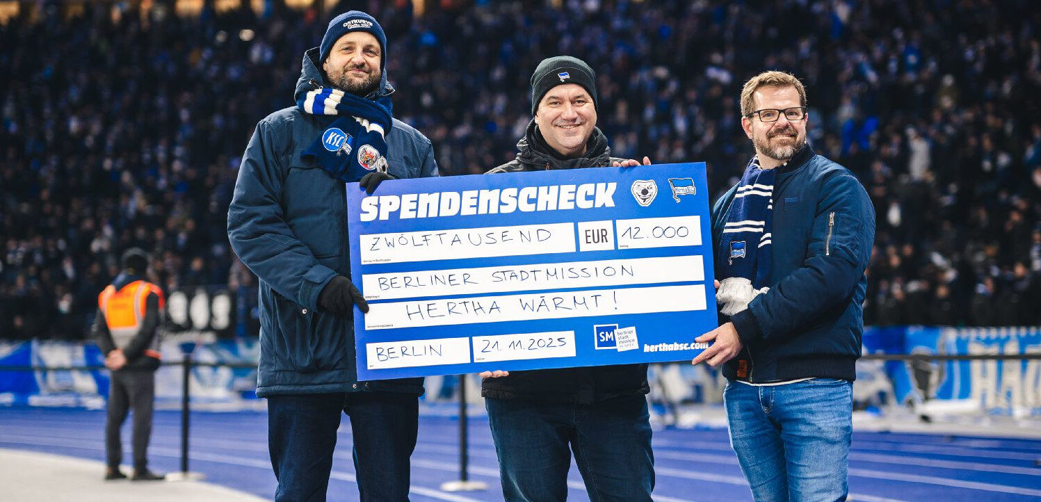 Drei Personen halten einen großen Scheck im Wert von 12.000 Euro für die Berliner Stadtmission im Rahmen der Aktion "Hertha wärmt!" auf einem Fußballfeld. Die Personen tragen blaue Kleidung mit Logos des Fußballvereins Hertha BSC. Im Hintergrund sind Zuschauer auf den Tribünen zu sehen.