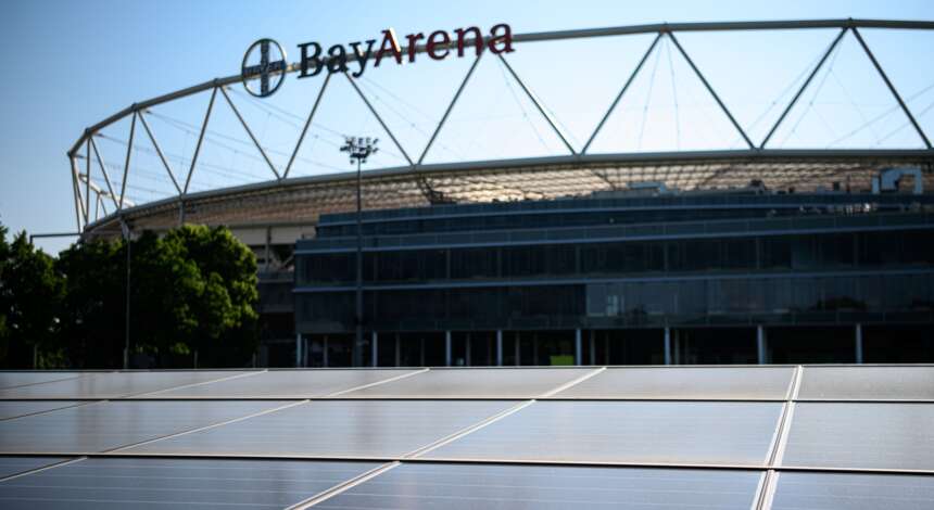 BayArena mit Solaranlage im Vordergrund. Das Stadion von Bayer 04 Leverkusen ist im Hintergrund zu sehen, mit dem Namen 'BayArena' prominent auf dem Dach. Die Solaranlage besteht aus mehreren Reihen von Solarpanelen, die vor dem Stadion platziert sind.