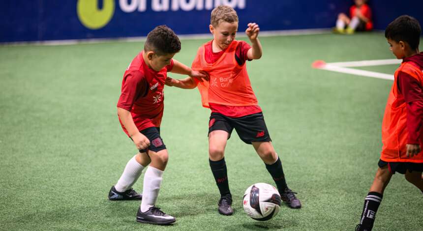 Zwei Kinder in roten Trikots spielen in einer Fußballschule in Leverkusen. Sie stehen auf einem grünen Kunstrasenfeld und versuchen, einen Fußball zu kontrollieren. Im Hintergrund sind weitere Kinder und eine blaue Wand mit einem Logo zu sehen.