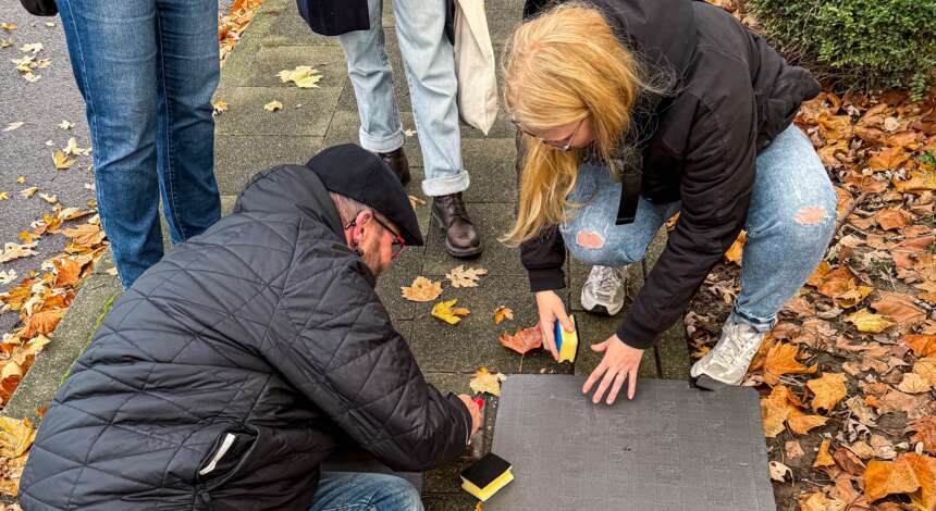 Zwei Personen knien auf einem Gehweg und setzen gemeinsam einen Stolperstein ein. Sie verwenden Werkzeuge, um den Stein in den Boden zu fixieren. Im Hintergrund sind weitere Personen zu sehen, die das Geschehen beobachten. Der Boden ist mit Herbstlaub bedeckt.