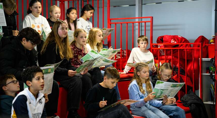 Eine Gruppe von Kindern und Jugendlichen sitzt auf roten Tribünenstühlen in einer Sporthalle und liest konzentriert in Broschüren oder Heften. Die Atmosphäre wirkt lernorientiert und aufmerksam.