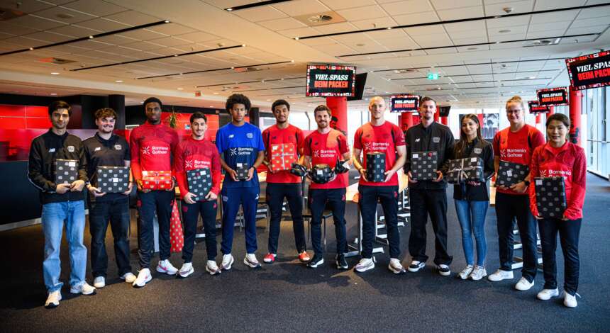 Eine Gruppe von Menschen, darunter Spieler und Mitarbeiter von Bayer 04 Leverkusen, stehen in einem Flughafen und halten jeweils einen schwarzen Koffer in den Händen. Sie tragen T-Shirts mit dem Aufdruck "Bayer 04 Leverkusen" und "Nachhaltigkeit". Die Gruppe posiert für ein Gruppenfoto vor einem Check-in-Bereich mit Schildern im Hintergrund.