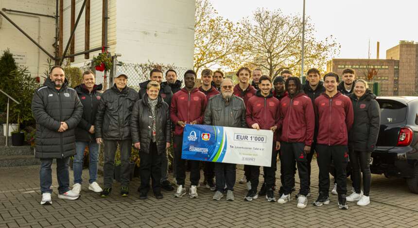 Gruppenfoto der U19-Mannschaft von Bayer 04 Leverkusen mit Vertretern der Leverkusener Tafel. Die Spieler und Begleitpersonen stehen vor einem Gebäude und halten einen Scheck über 1000 Euro sowie einen Gutschein über Lebensmittel im Wert von 500 Euro. Alle tragen warme Jacken und lächeln in die Kamera.