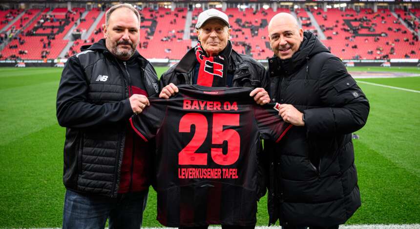 Drei Männer stehen auf einem Fußballfeld und halten gemeinsam ein Trikot mit der Aufschrift "Bayer 04 25 Leverkusener Tafel". Sie lächeln in die Kamera und tragen warme Kleidung, darunter Jacken und ein Schal mit dem Vereinslogo. Im Hintergrund sind die roten Sitze des Stadions zu sehen.